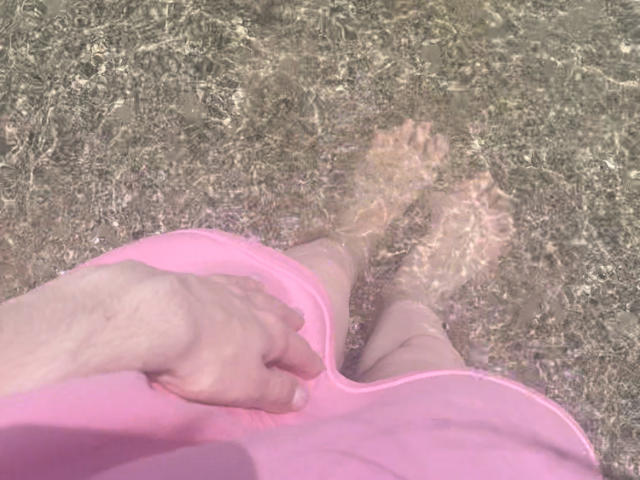 A calça rosa de RozalinaPleasure, com a mão apoiada em suas nádegas, pés descalços em água cristalina, a luz do sol refletindo em seus dedos. Sua calça rosa contrasta com os reflexos claros da água..