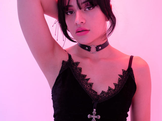 DaliaBelle con un top de encaje negro, un collar choker negro con un anillo, un piercing en la nariz y el brazo levantado, mostrando su axila. Tiene una iluminación rosa en su piel. Sus pechos son pequeños..
