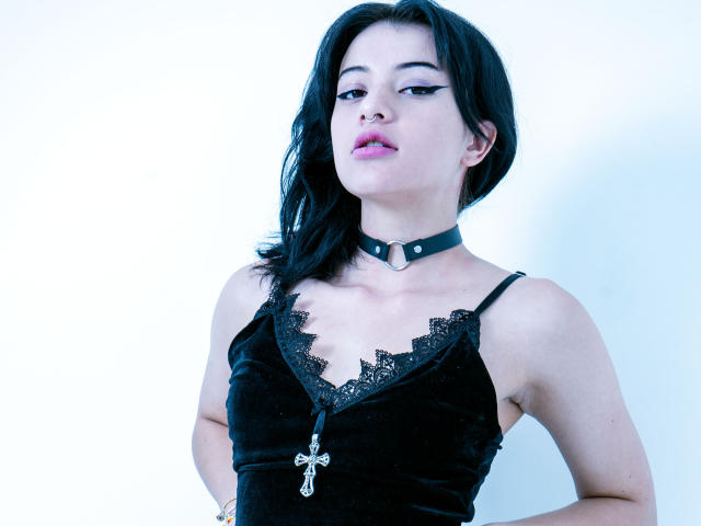 DaliaBelle, con cabello negro, vestida con una blusa de terciopelo negro con encaje, un collar negro y un collar con una cruz, con las manos en las caderas, mirando ligeramente hacia arriba, labios de color rosa pálido..