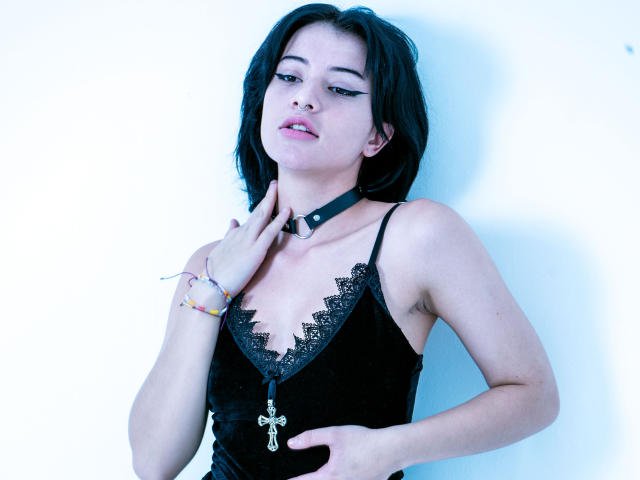 DaliaBelle portant un caraco en dentelle noire, un collier ras du cou noir, une croix en pendentif et un bracelet coloré, une main sur la poitrine, l'air légèrement agacée, peau pâle, cheveux foncés, photo avec une teinte bleutée..