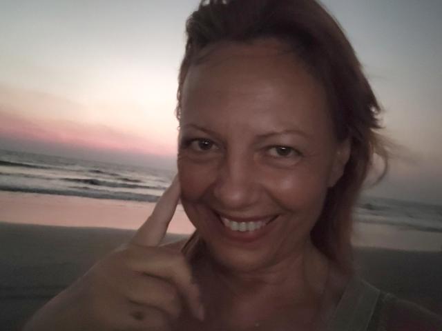BarbaraBowie, smiling with her finger touching her cheek, has light brown hair, is wearing a green top, and is at a beach during sunset. Her tits are partially visible.