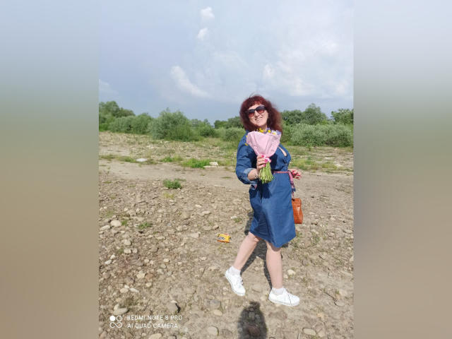 BellisimaAh mit lockigem, rotem Haar, trägt eine Sonnenbrille, ein blaues Kleid, weiße Turnschuhe, hält einen pinken Schal und eine braune Tasche, steht auf felsigem Untergrund und lächelt. Ihre Brüste sind klein, ihr Hintern ist niedlich..
