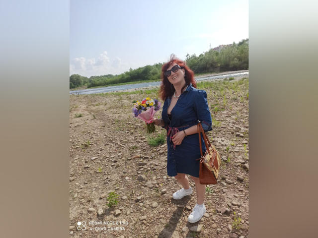 BellisimaAh met rood krullend haar, gekleed in een blauwe jurk, witte sneakers en een zonnebril, met een boeket in haar handen, staand op rotsachtig terrein, met een bruine tas over haar schouder, lachend..