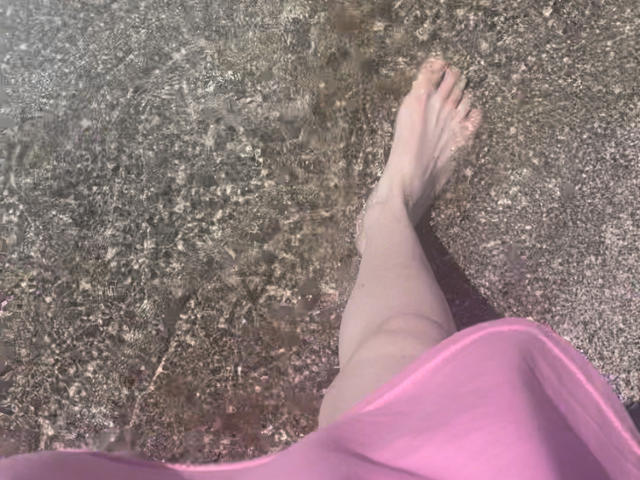 Rozalina's bleke been en voet in ondiep, helder water, met een roze stof die gedeeltelijk haar onderlichaam bedekt, staand op een ruwe, rotsachtige bodem..