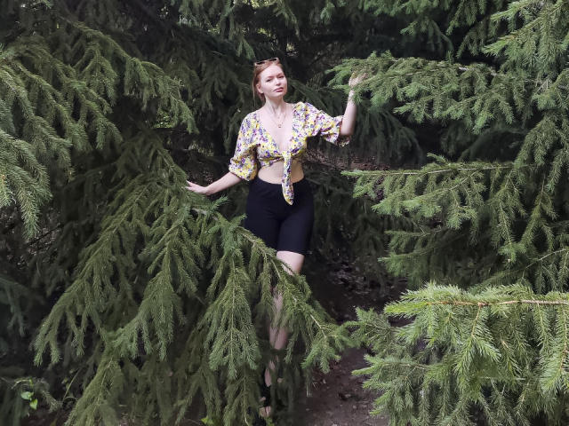 SweetAleksaX se tient au milieu de grands pins, vêtue d'une chemise à imprimé floral avec un nœud devant, de shorts de vélo noirs, et porte des lunettes de soleil sur la tête, avec sa main posée sur une branche de pin..