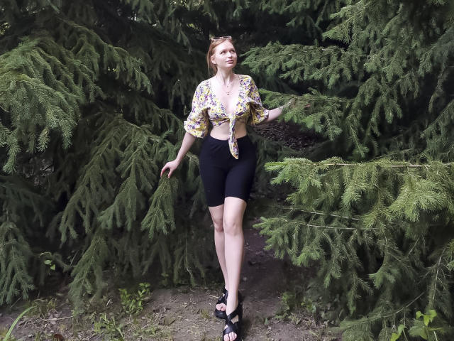 SweetAleksaX with tied-up floral blouse, black bike shorts, and black heels, standing among green pine trees, touching branches with right hand, looking up, blonde hair, fair skin.