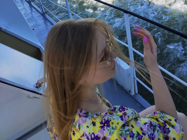 SweetAleksaX met blond haar, draagt een bloemenjurk, haar in handen, roze nagels, boot, zon die reflecteert in de bril, water op de achtergrond, kijkt naar de zijkant, laat borsten zien, mooi, stijlvol..