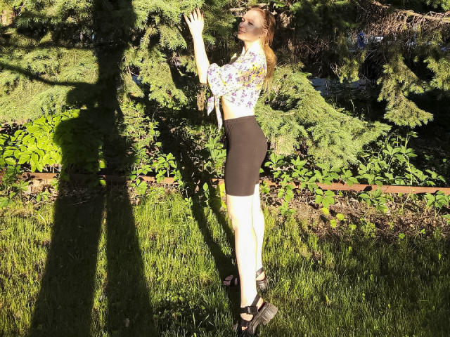 SweetAleksaX stands in sunlight, reaching up, wearing a floral blouse, black skirt, and black heels, with her ass slightly raised, shadowed on green grass and pine trees.