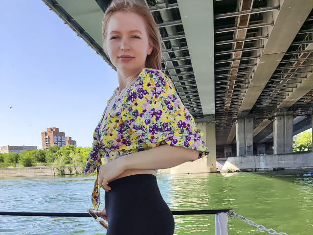 SweetAleksaX staat langs een groene rivier, gekleed in een geel bloemenblousje dat aan de voorkant is vastgebonden en zwarte broeken met hoge taille, onder een brug met een metalen dak..