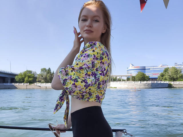 SweetAleksaX with red hair, wearing a yellow floral top tied at the front and black high-waisted pants, standing on a boat, looking over her shoulder, holding her chin.