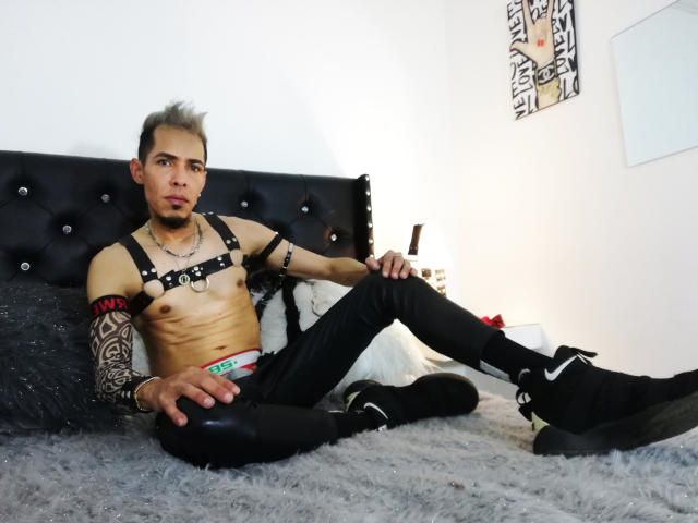 JeikkoHot, ohne Hemd, mit schwarzem Ledergeschirr, Tätowierungen am rechten Arm, sitzt auf dem Bett mit gespreizten Beinen, trägt schwarze Hosen, Nike-Sneakers, graues, stacheliges Haar, blickt in die Kamera und hält sich selbst..
