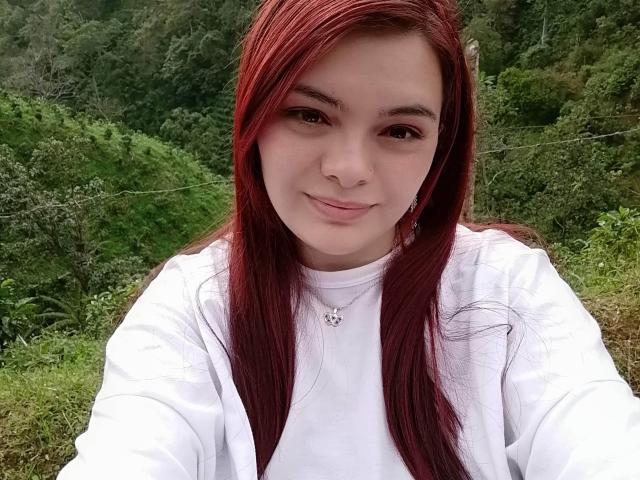 KatherinLatin ha i capelli rossi, una camicia bianca e un sorriso delicato, con un piccolo piercing al naso e una collana. Il suo seno non è visibile. È all'aperto e sembra molto carina..