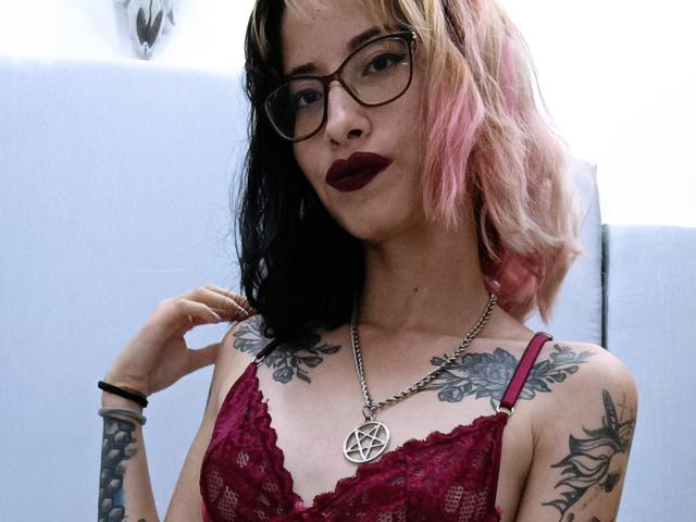 LailaStewart with pink and black hair, glasses, dark lipstick, red lace bra, tattoos on shoulders and forearm, pentagram necklace, hand near head, looking at camera.