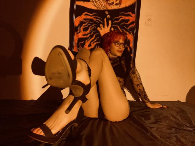 LailaStewart with red hair, glasses, and black lace top, sitting on bed, legs up, showing black high heels, ass, and tattooed arm, in warm, dim lighting.