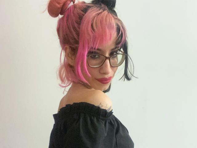 Laila Stewart, met roze en zwarte haren, een bril en een zwarte top met een open schouder, kijkt over haar schouder, waarbij een tatoeage op haar rechter schouder zichtbaar is, draagt roze lippenstift en glimlacht..