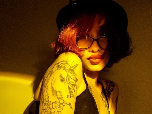 Laila Stewart, met rood geverfd haar, een zwarte hoed, een bril en tatoeages op haar arm en borst, laat haar borsten zien in een zwarte top, verlicht door geel licht..