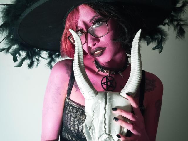 Laila Stewart, con piel rosada, cabello rojo y negro, gafas, blusa negra de encaje, collar negro, pentagrama, cráneo con cuernos y un gran sombrero negro de bruja, sosteniendo cuernos, con esmalte de uñas negro..