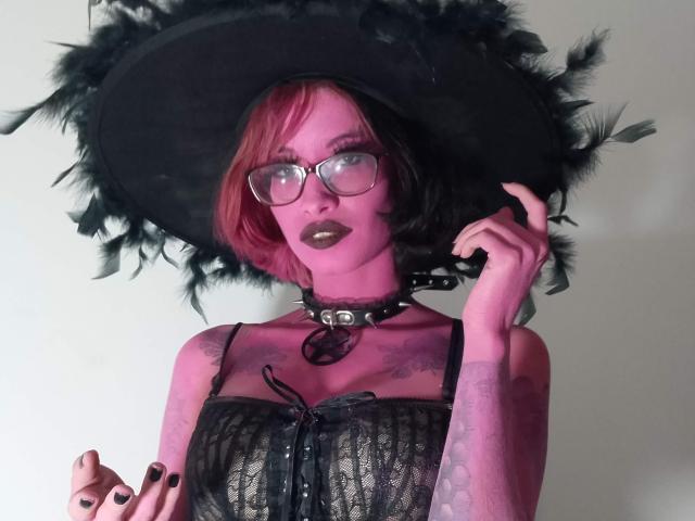 Laila Stewart, met roze en zwarte gekleurd haar, draagt een bril, een zwarte kanten jurk en een zwarte hoed met veren. Ze houdt de hoed vast, draagt een choker met een ring en heeft tatoeages op haar armen. Ze heeft donkere lipstick op..