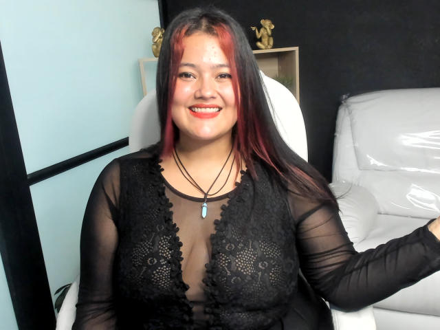 VaneParker tem cabelo ruivo e preto, um sorriso adorável, e veste uma blusa preta de renda transparente que revela seus seios. Ela está sentada em uma cadeira branca, sorrindo..