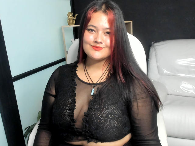VaneParker con reggiseno a fascia in pizzo nero, maniche trasparenti, riflessi rossi nei capelli neri, collana, sedia bianca, divano bianco, sorridente alla fotocamera, che mostra il suo seno prosperoso..