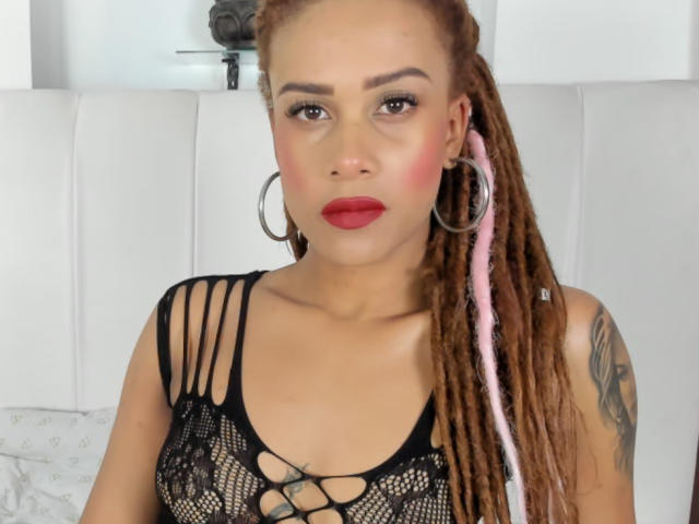 KendallQuinn has light brown skin, red lipstick, and pink-and-brown braided hair. She's wearing a black, lace-up, see-through top, showing her small tits and a tattoo on her right arm.
