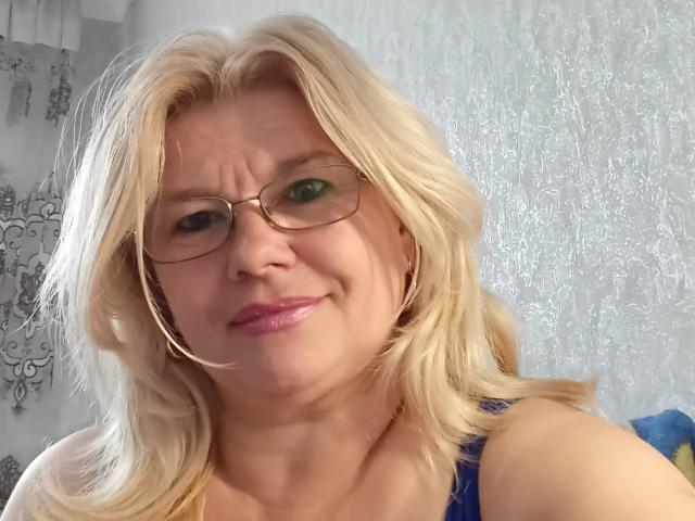 DreamyShirley, com cabelo loiro, óculos, batom rosa, sorrindo levemente, mostrando seios pequenos, usando uma blusa azul, em um selfie. Ela é uma mulher branca de meia-idade com olhos verdes..