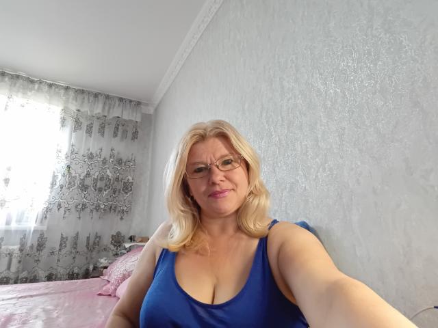 Blond, verträumte Shirley mit Brille, trägt ein blaues Tanktop, zeigt ihre großen Brüste, sitzt auf einem rosa Bett und lächelt in die Kamera..