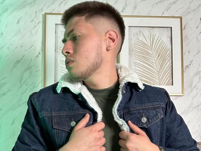 SebastiianMaxime with short brown hair, slight beard, looking to the side, wearing a blue denim jacket with white fleece lining, hands on jacket, framed leaf art in background.