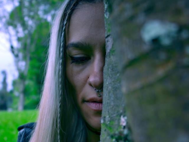 Amelie Blake, con i capelli biondo platino, un piercing al naso e l'eyeliner scuro, parzialmente nascosta da un tronco d'albero, mostra un'espressione calma e seria, con uno sfondo verde sfocato..
