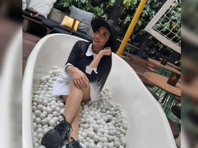 SarahyJhones sitting in a white bathtub filled with white pebbles, wearing a black hat, black shirt with white collar, white skirt, and black sneakers, looking to the side.