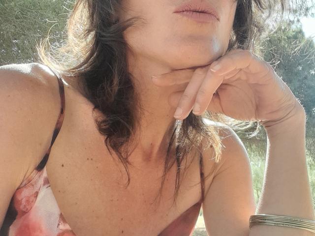 Le visage d'Aiyana est partiellement visible, avec des cheveux bruns ondulés et des lèvres roses, le menton reposant sur sa main. Elle porte une robe fleurie rose et rouge, baignée de soleil..