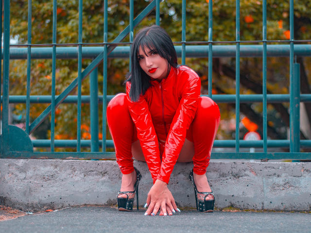 Giuliana Fetich in posizione accovacciata, indossa un completo in lattice rosso lucido, tacchi alti neri, rossetto rosso, taglio di capelli corto nero, mani appoggiate sul marciapiede, una recinzione in metallo verde sullo sfondo, foglie autunnali visibili..