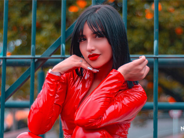 Giuliana Fetich vestida con látex rojo brillante, cabello negro, lápiz labial rojo, brazos cruzados, mano en la barbilla, sonriendo, cerca hay una valla de metal verde, luciendo sexy con un "escote" sutilmente insinuado..