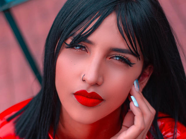 GiulianaFetich tem cabelo preto, olhos azuis, batom vermelho, um piercing no nariz e unhas azuis, vestindo uma blusa vermelha brilhante e olhando sedutivamente para a câmera..
