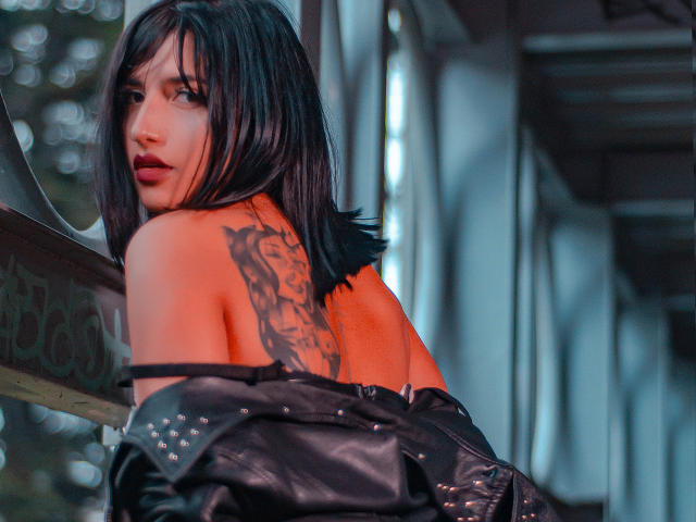 GiulianaFetich with black hair, red lipstick, and a detailed tattoo on her back, wearing a black leather jacket, looking over her shoulder. Her ass is partially visible.