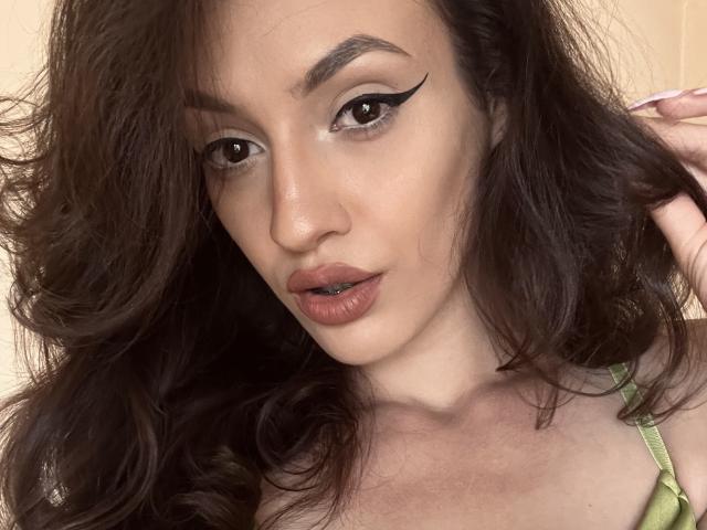 VievienneClemente with dark, wavy hair, thick black eyeliner, and full pink lips, showing a subtle pout. She's wearing a green top, with a hand in her hair.