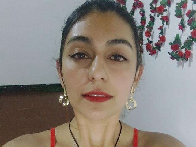 Rosalie Black hat eine mittelbraune Haut, dunkles Haar zu einem Dutt gebunden, roten Lippenstift, schwarzen Eyeliner, goldene Ohrringe, ein rotes Oberteil und eine schwarze Halskette und blickt direkt in die Kamera..