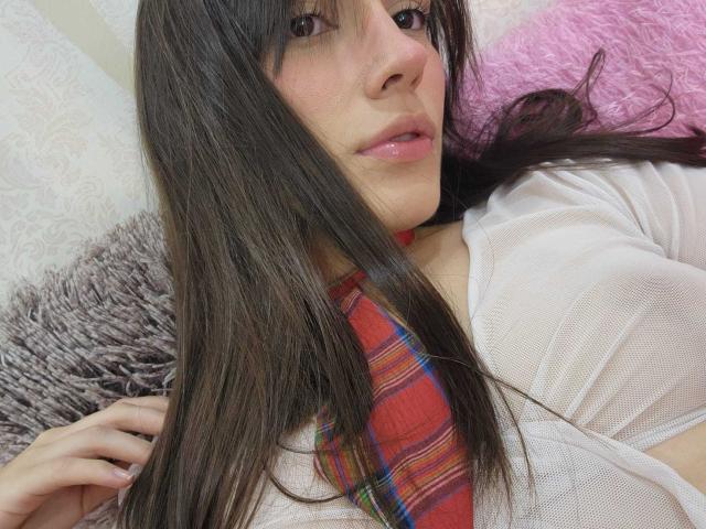 MellyDolly con cabello largo y castaño, labios rosados, vistiendo una blusa blanca transparente y una corbata de cuadros rojos, acostada sobre un cojín rosa, con la mano en el cabello, sonriendo ligeramente, mostrando pequeños senos..