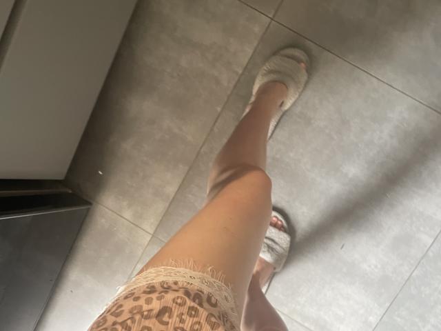 RozalinaPleasure's been heeft haar been omhoog gestrekt, draagt witte sneakers en ondergoed met een kantpatroon, waarbij haar blote dij en witte sokken zichtbaar zijn, met een grijze betegelde vloer op de achtergrond..