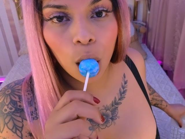 Chili chupa um pirulito azul, os olhos meio fechados, ombros tatuados visíveis, esmalte vermelho na mão direita.