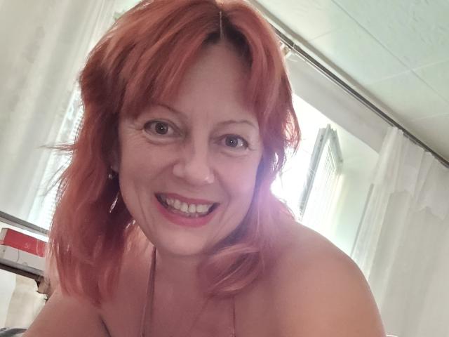 BarbaraBowie with red hair, smiling, showing her tits, wearing a halter top, in a selfie with bright light behind her.
