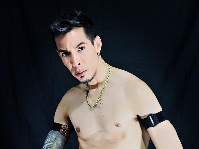 Shirtless JeikkoHot with short black hair, chain necklace, black arm bands, tattoo on right arm, and ear studs, looking down with a slight smile. Dark background.