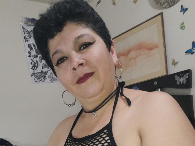 Angellovelly has short black hair, dark makeup, red lipstick, hoop earrings, black fishnet top, choker with a heart, and is smiling at the camera with her tits partially visible.