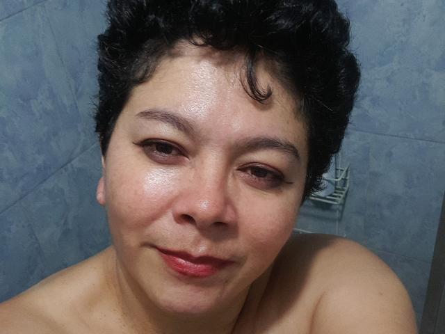 Angellovelly, con cabello negro rizado y corto, piel ligeramente húmeda y una sonrisa sutil, mirando a la cámara con lápiz labial rojo. Hombros desnudos visibles..