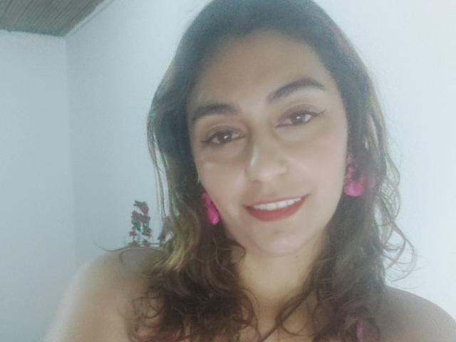 RosalieBlack with wavy brown hair, pink earrings, and red lipstick smiling at the camera, showing her cute face and small tits. She's wearing a pink top, looking cute.