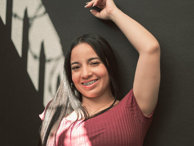 Gianna Russell, avec de longs cheveux noirs, souriante, bras levé, portant un t-shirt rose côtelé, montrant des bagues dentaires métalliques, debout devant un mur sombre et texturé..