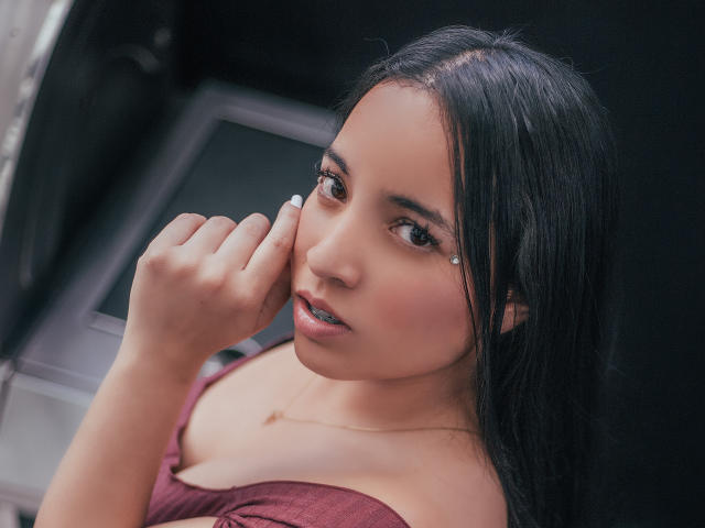 Gianna Russell, con cabello negro, ojos marrones y una lágrima en su mejilla, tocándose la cara, vistiendo una blusa color burdeos, mostrando sus senos, y un collar pequeño..