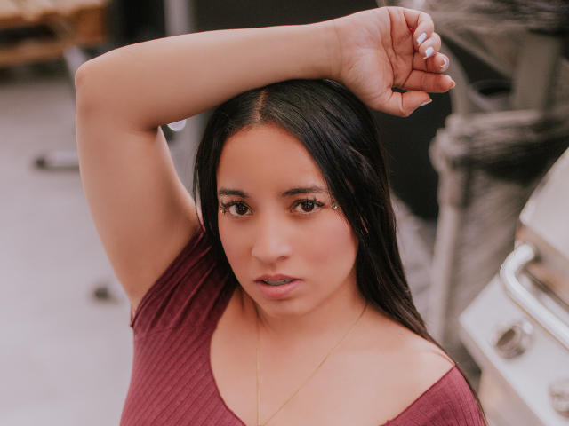 Gianna Russell, con cabello largo y negro, levantando su brazo derecho, vistiendo una camiseta de color burdeos con textura acanalada y un collar dorado, mirando directamente a la cámara con una expresión neutra..
