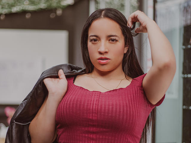 Gianna Russell, con cabello negro largo, vestida con una camiseta roja texturizada, mostrando sus senos, sosteniendo una chaqueta negra, con expresión seria, brazo derecho levantado, un piercing discreto en la nariz, collar de oro..
