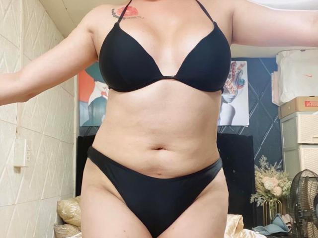 TsCockyLiciousz trägt einen schwarzen Bikini, zeigt ihre Brüste und ihren Hintern, hat ein Tattoo an der rechten Schulter und einen leicht dicken Bauch und steht in einem Raum..